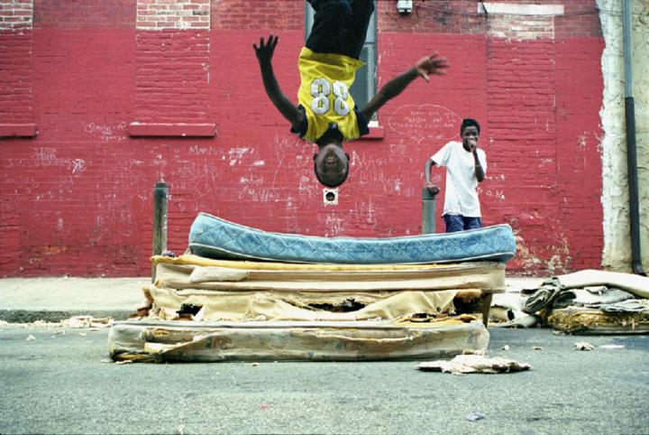 A young black boy backflipping onto a blown out mattress in the middle of the street.