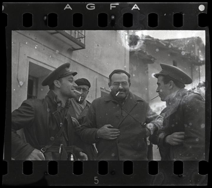 Soldiers laughing in a circle with unlit cigarettes, fishing in their pockets for one. 